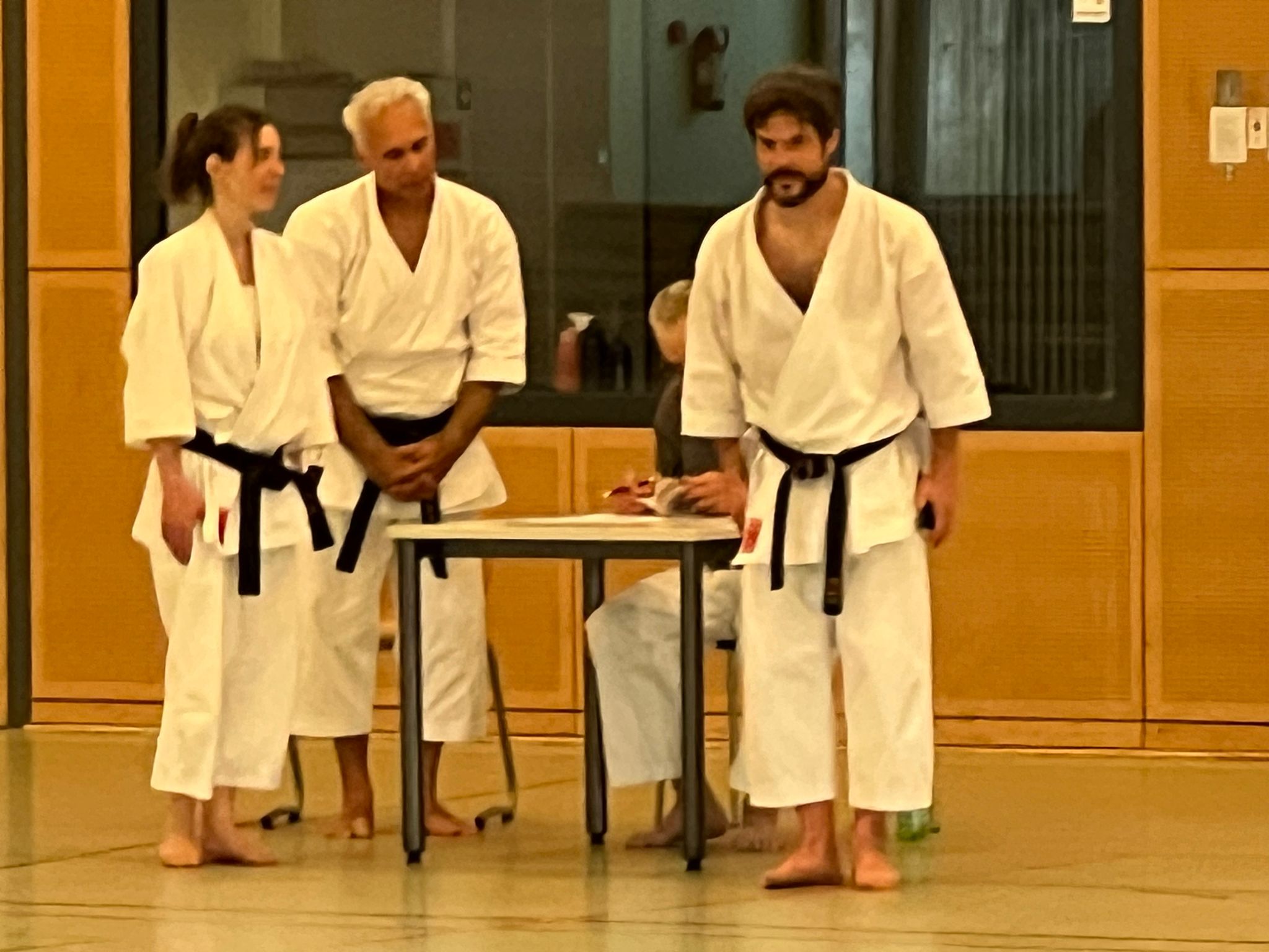 Drei Personen in weißen Karate-Gis stehen an einem Tisch, konzentrierte Gesichter, Holzvertäfelung.
