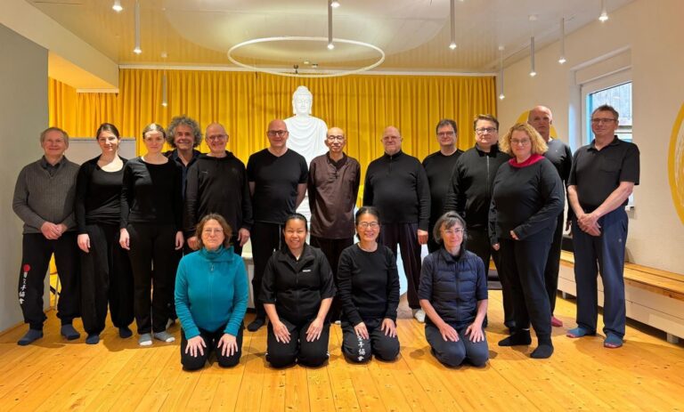 Gruppe von Menschen in dunkler Kleidung vor gelben Vorhängen, lächelnd, mit Buddha-Statue im Hintergrund.