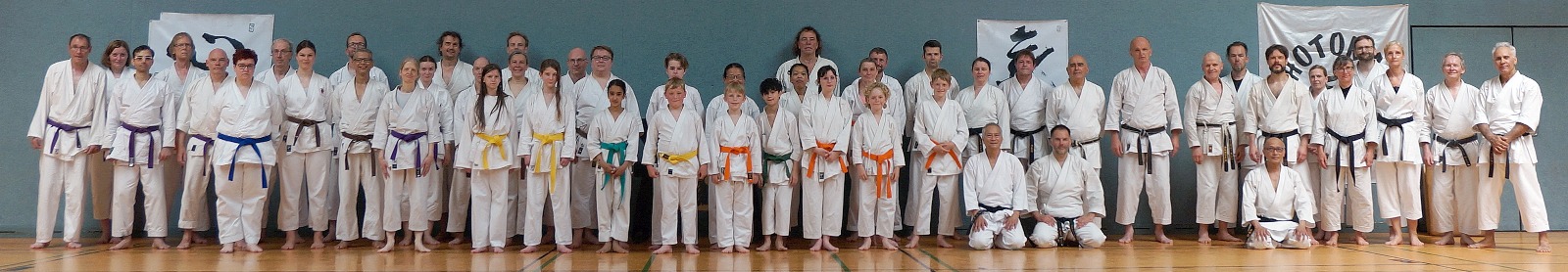 Gruppe von Menschen in Karate-Gis posiert lächelnd in einer Turnhalle mit bunten Gürtelstufen.