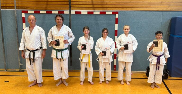 Mehrere Menschen in Karateanzügen zeigen Zertifikate, lächeln in einer Sporthalle.