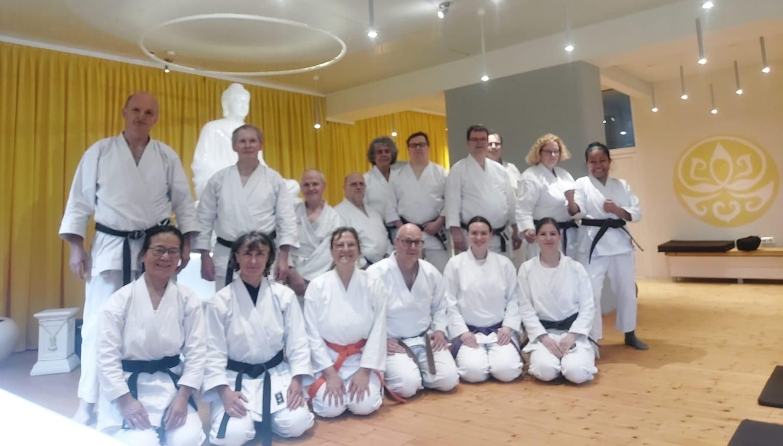Gruppe in Karate-Gi lächelt in hellem Raum mit gelben Vorhängen und Buddha-Statue im Hintergrund.