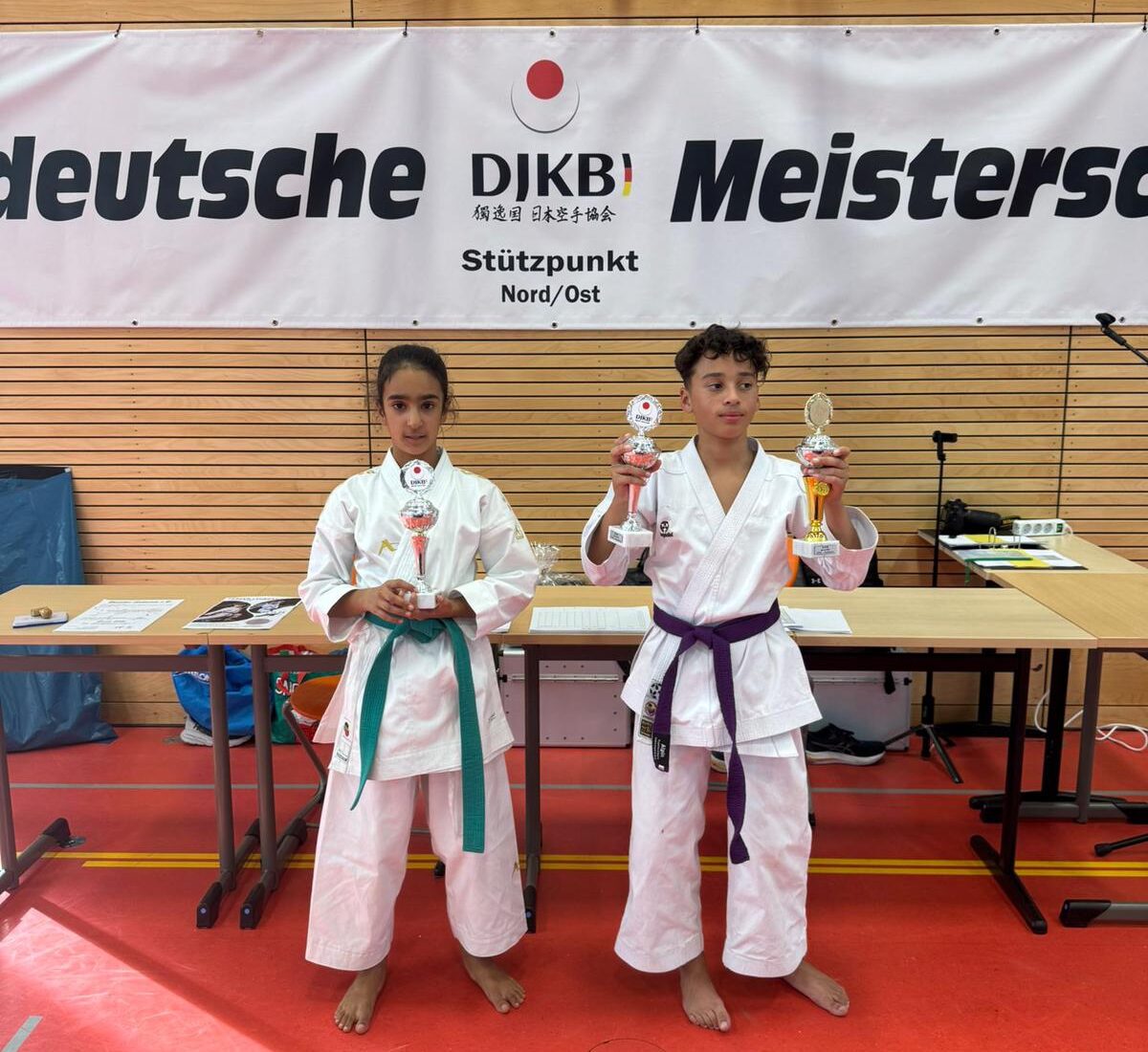 Zwei Kinder in Karateanzügen halten Trophäen, mit zufriedenen Gesichtsausdrücken vor einem Banner für eine deutsche Meisterschaft.