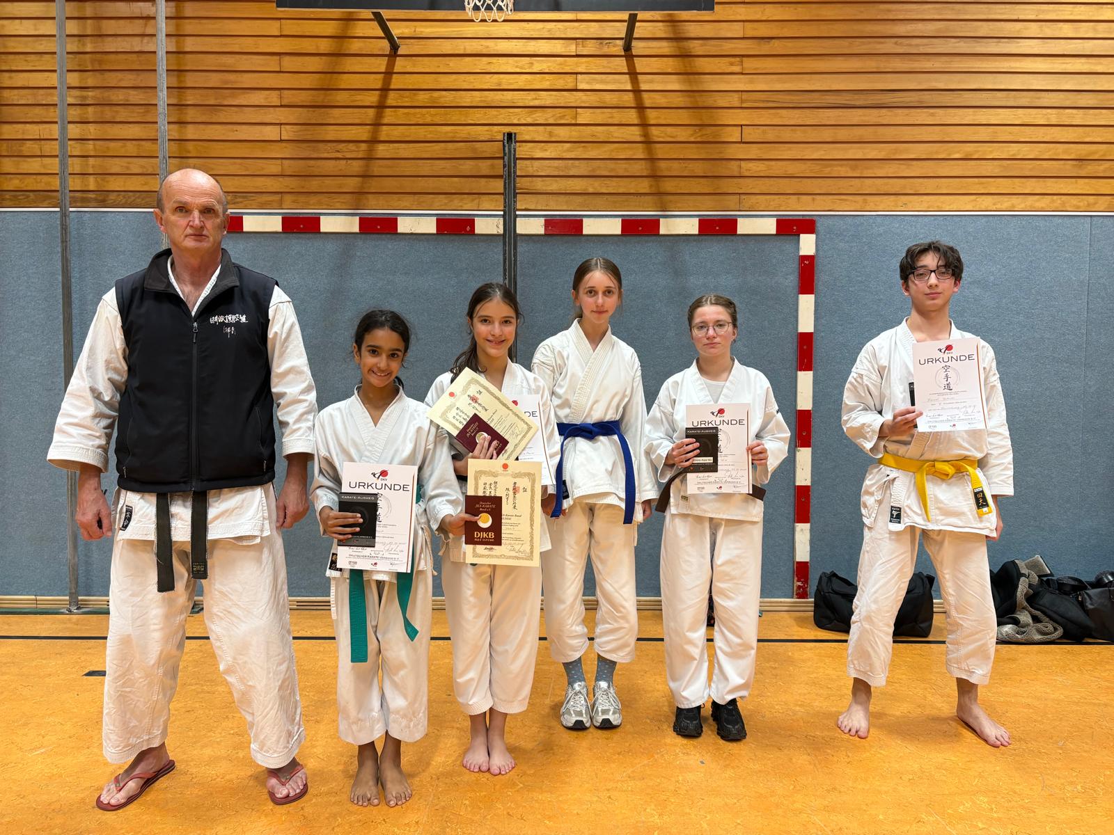 Gruppe von sechs Personen in Karateanzügen, vier Kinder halten Urkunden, alle lächeln in einer Sporthalle.