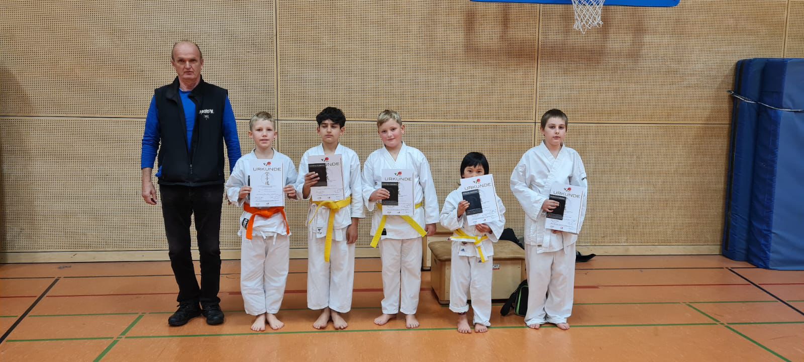 Eine Gruppe von fünf Kindern in weißen Karateanzügen hält Urkunden, mit einem erwachsenen Mann in schwarzer Kleidung daneben in einer Turnhalle.