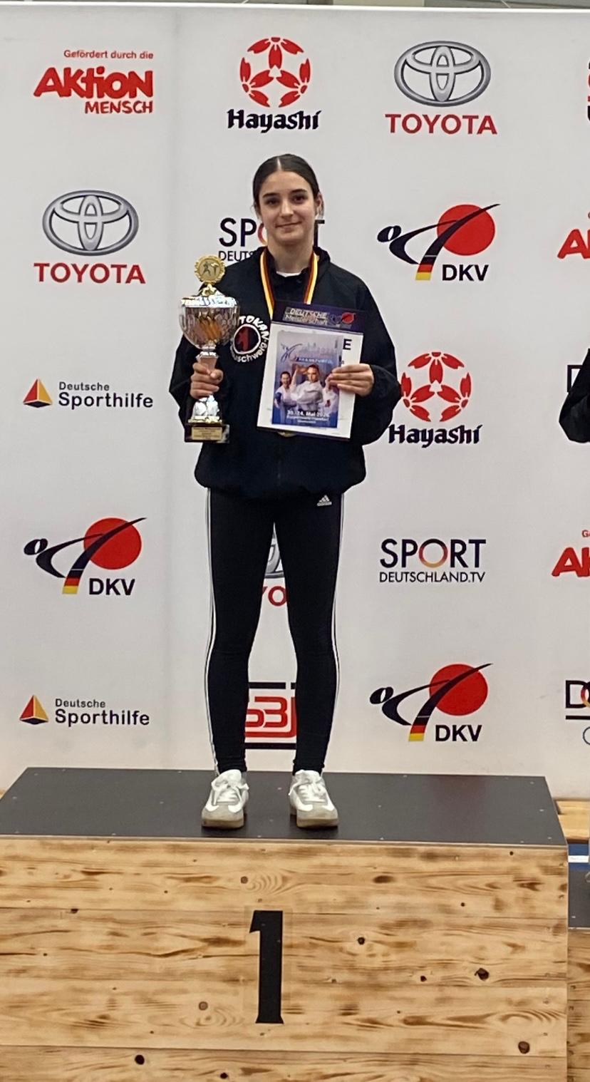 Junge Frau mit dunklem Haar lächelt stolz auf dem Siegerpodest mit Pokal und Urkunde