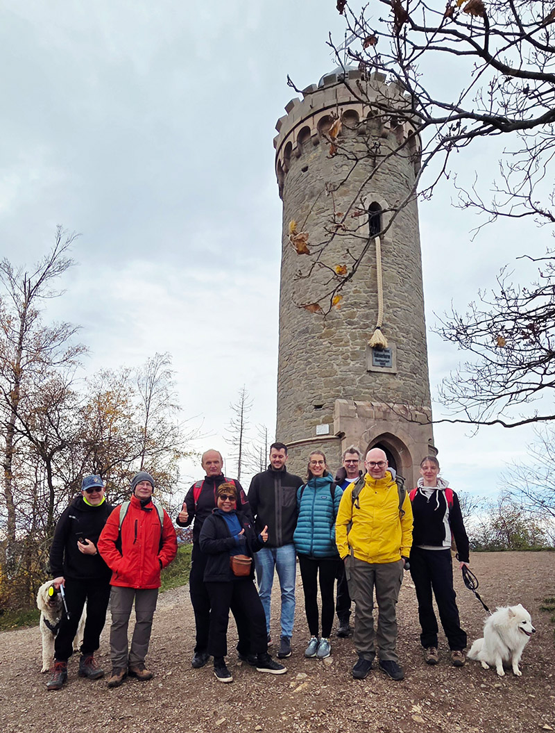 Neun lächelnde Menschen mit bunten Jacken stehen vor einem historischen Steinturm