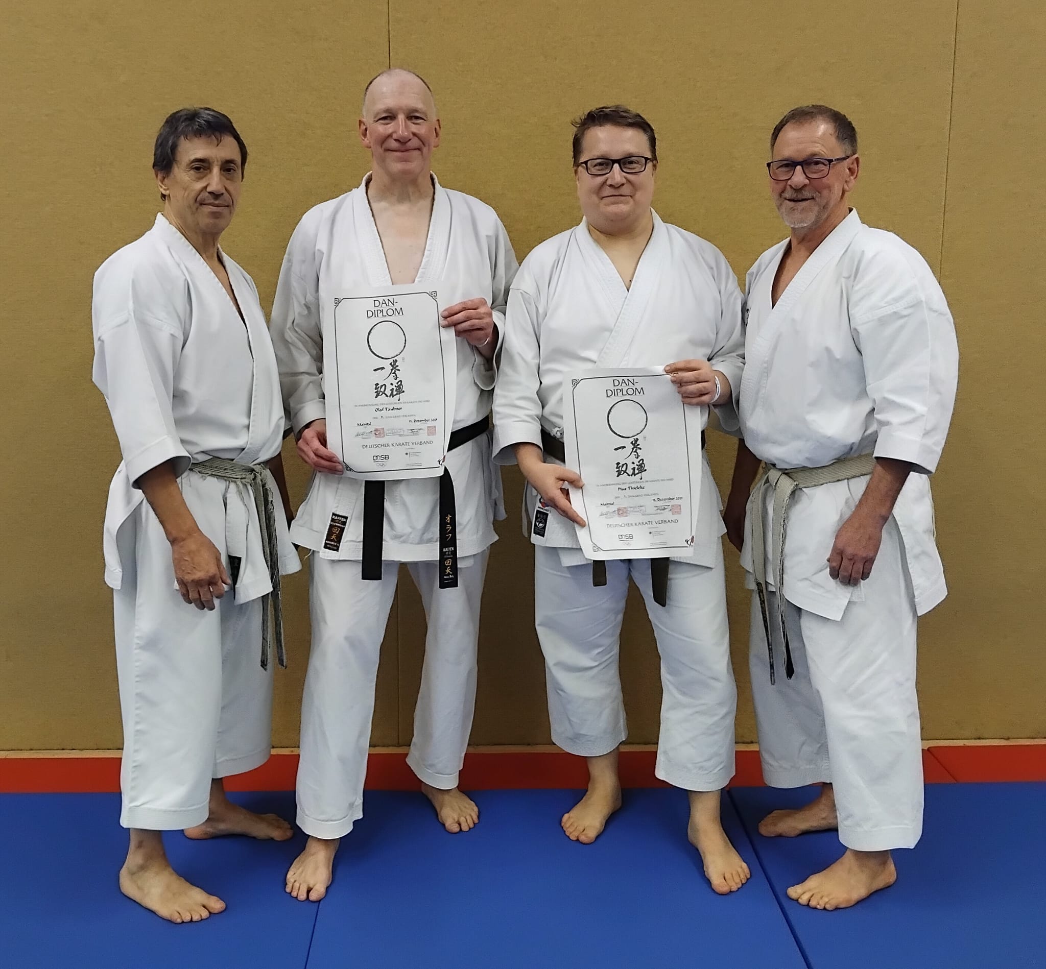 Vier Männer in weißen Karateanzügen lächeln, zwei halten stolz Urkunden hoch
