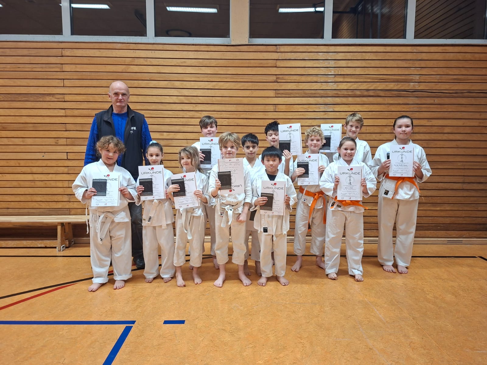 Eine Gruppe von Kindern im weißen Karateanzug lächelt stolz mit Urkunden in der Halle
