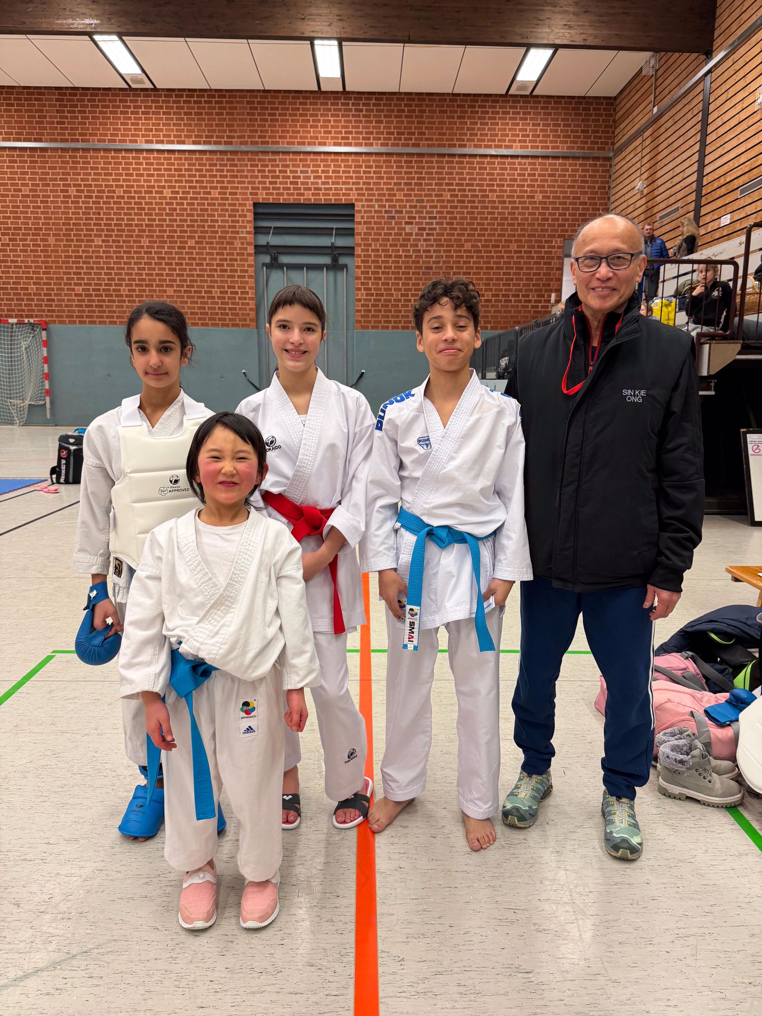 Vier Kinder in Karateanzügen und ein älterer Mann lächeln gemeinsam in einer Sporthalle
