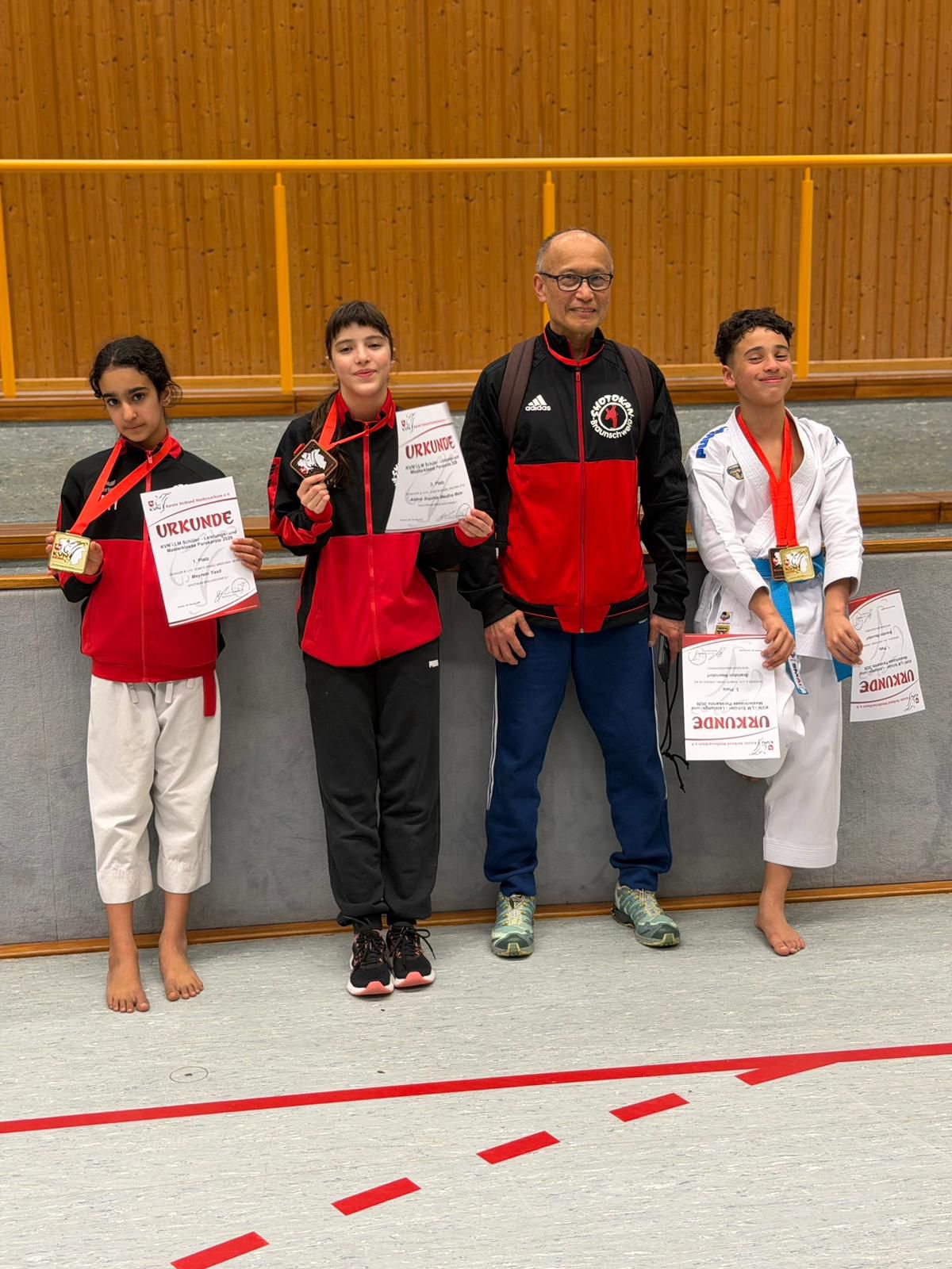 Drei Jugendliche und ein älterer Mann posieren stolz mit Urkunden und Medaillen in rotweißer Sportkleidung vor einer Holzverkleidung