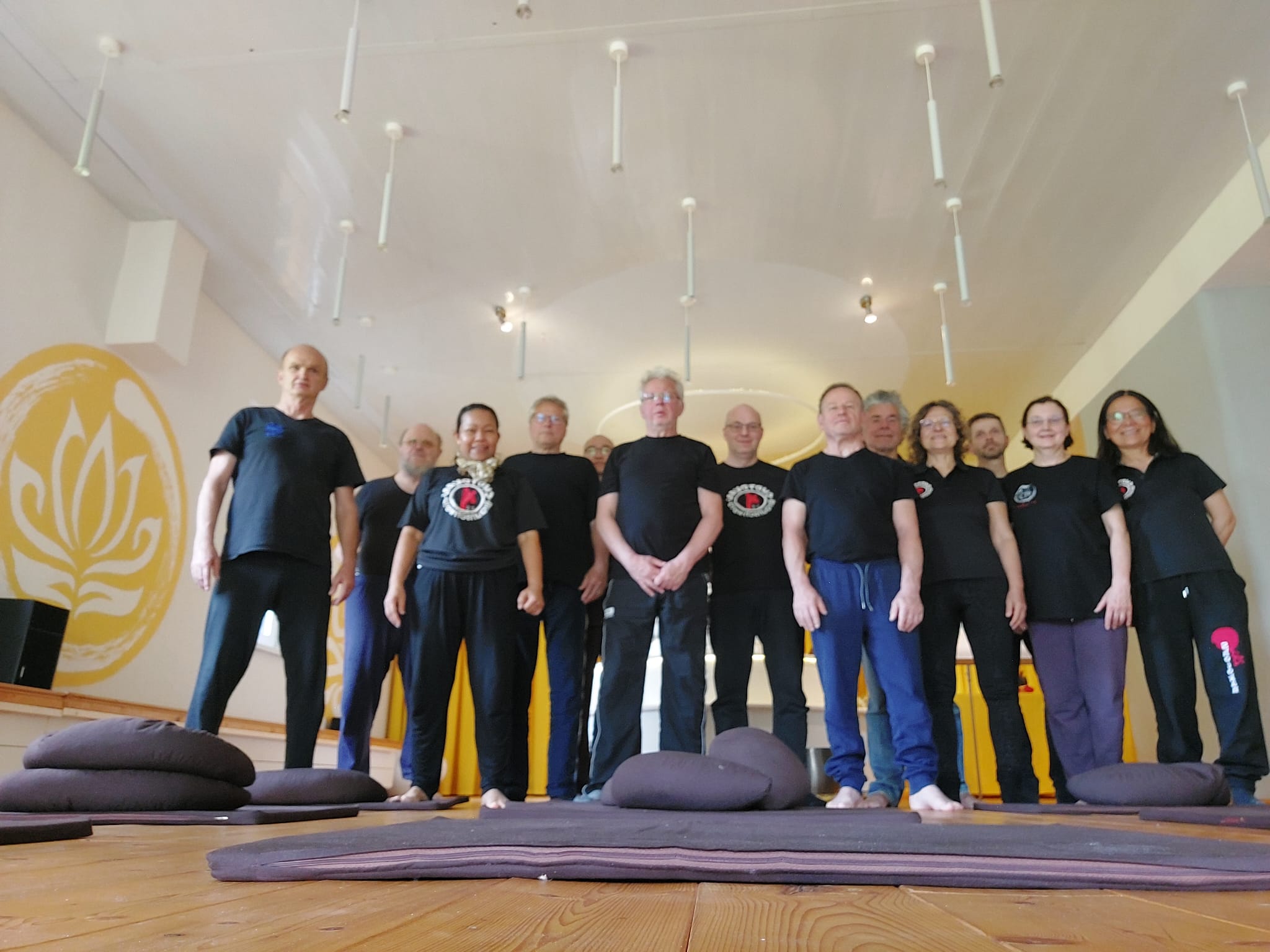 Eine Gruppe von Menschen steht lächelnd in schwarzer Kleidung in einem hellen Yogaraum