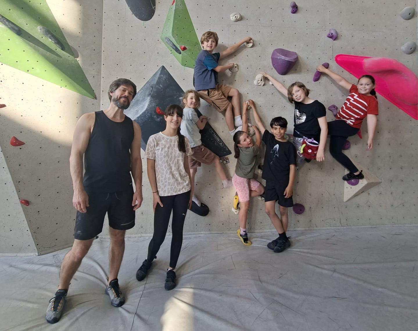 Mehrere lachende Kinder und zwei Erwachsene an grauer Kletterwand mit bunten Griffen