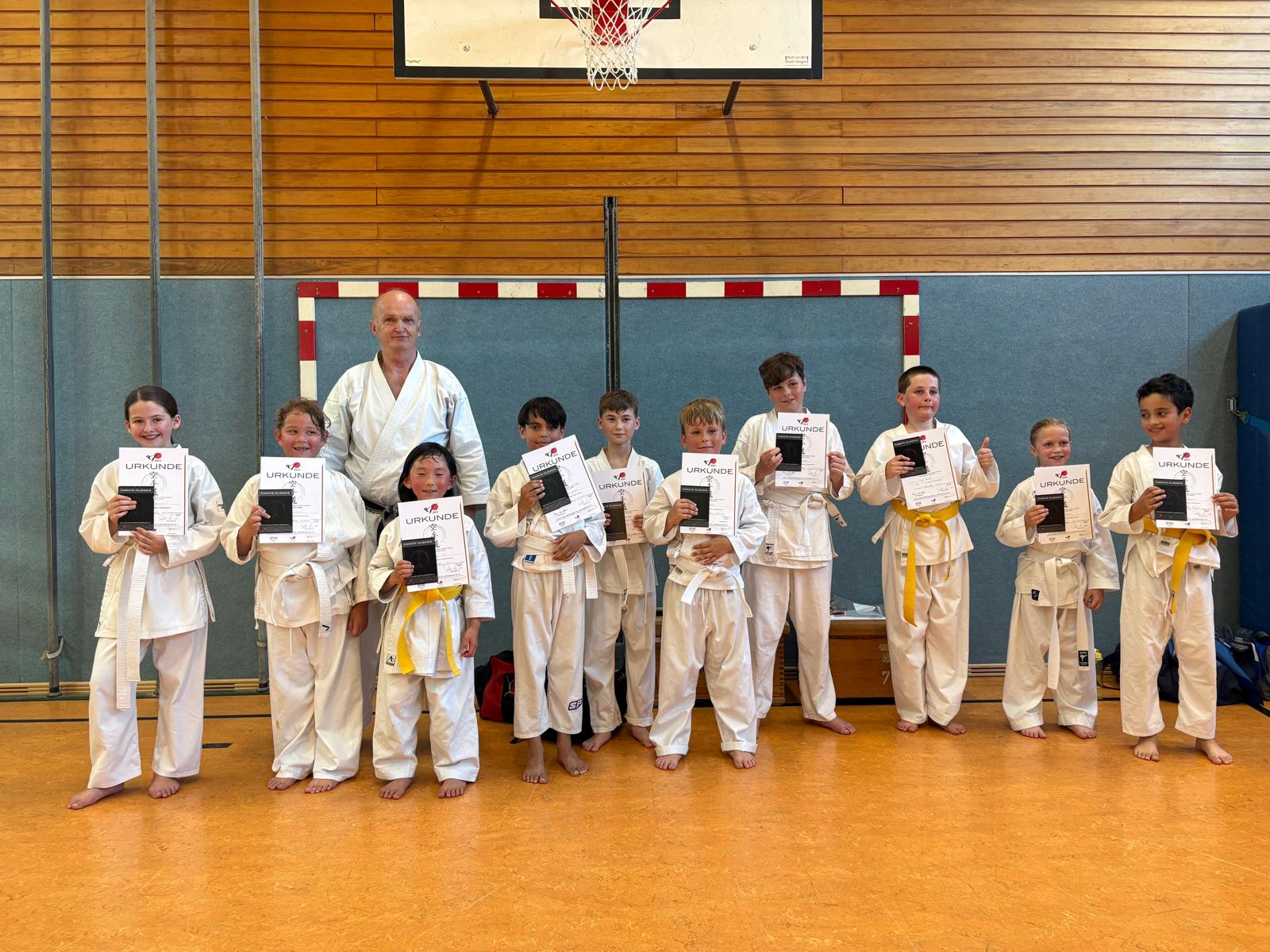 Kinder in weissen Karateanzuegen lachen und halten Urkunden in Turnhalle