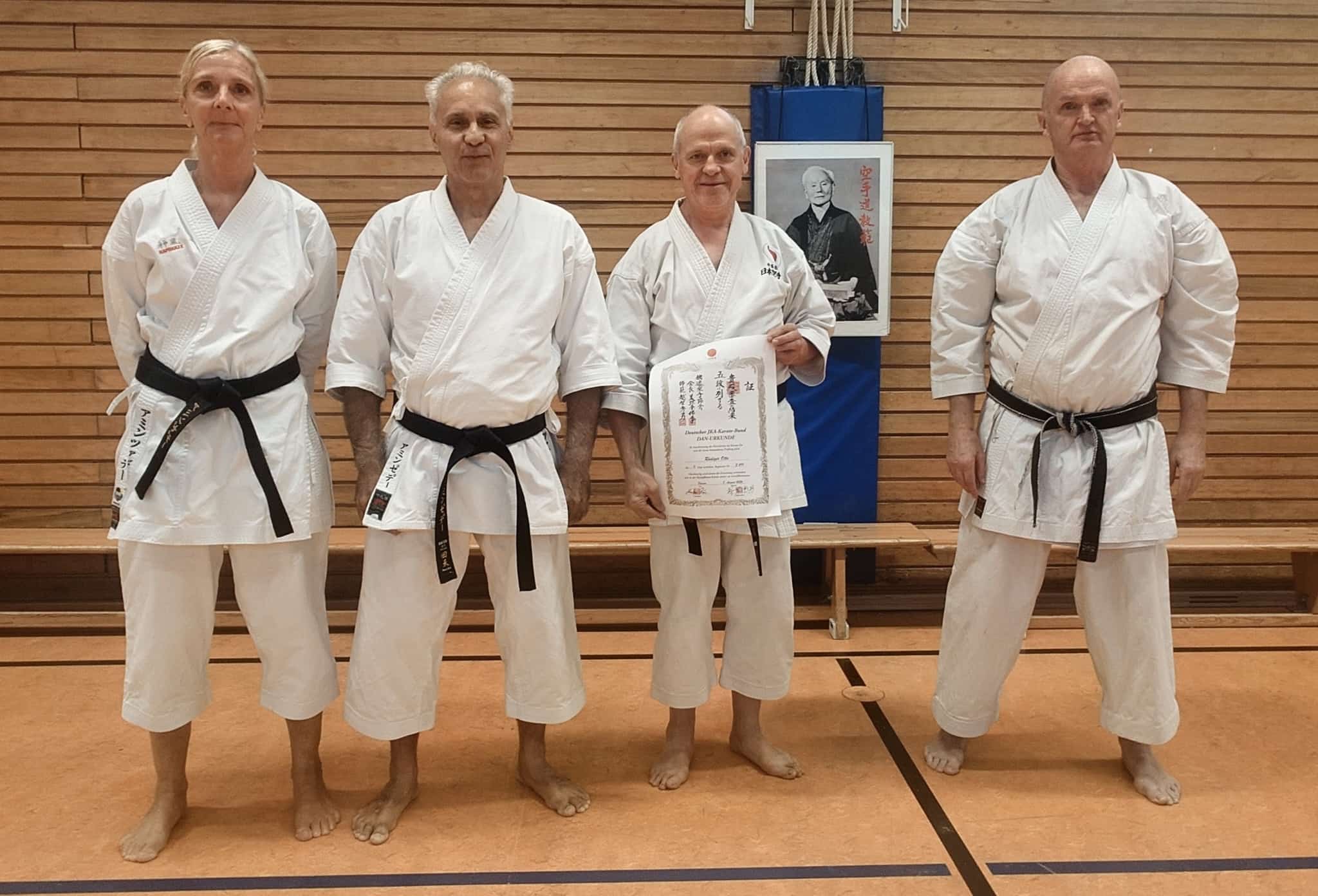 Vier laechelnde Karatekas in weissen Anzuegen mit schwarzen Guerteln und einer Urkunde